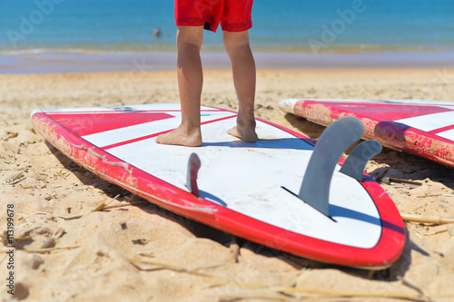 Fun activity on surf camp vacation. Back view of small boy young surfer with surfboard on sand beach.