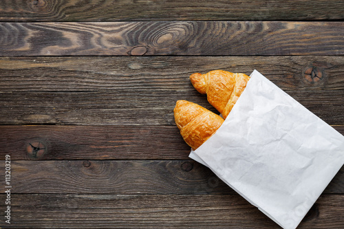 Tasty croissants in white paper bag. Rustic wooden background with sweet dessert. Top view.