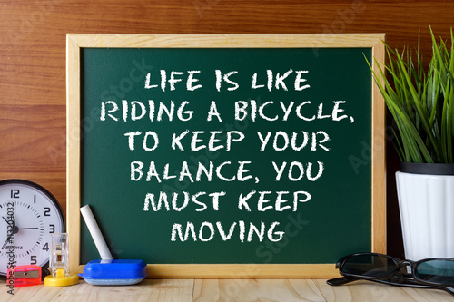 Word quote LIFE IS LIKE RIDING A BICYCLE,TO KEEP YOUR BALANCE,YOU MUST KEEP MOVING written on green chalk board on wooden table.