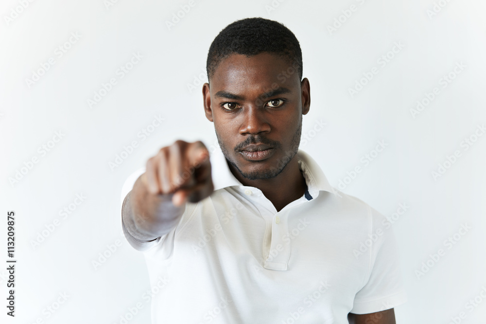 Headshot of African man wearing white polo shirt, pointing a finger at the camera with serious expression, saying that he is choosing you, against white studio wall background. Selective focus Headshot of African man wearing white polo shirt, pointing a finger at the camera with serious expression, saying that he is choosing you, against white studio wall background. Selective focus