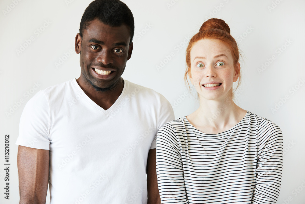 A couple grinning and making scary faces with false smiles and wide eyes. African man staring at camera and red-head Caucasian girl is freezed in stupefaction. Strange and crippy mimics, fun concept. A couple grinning and making scary faces with false smiles and wide eyes. African man staring at camera and red-head Caucasian girl is freezed in stupefaction. Strange and crippy mimics, fun concept.