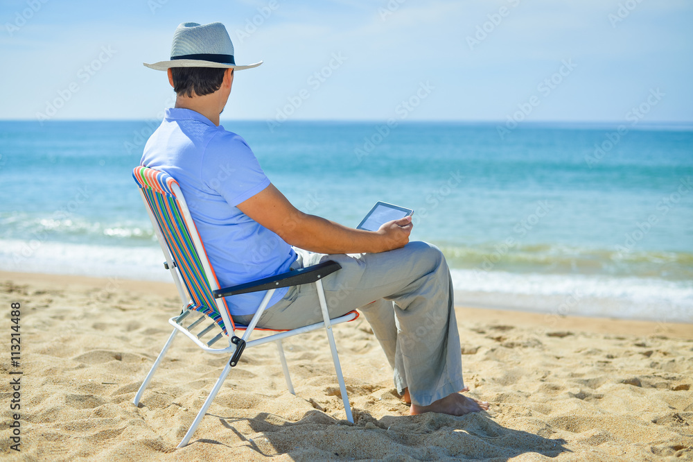 Man using tablet relaxing on ocean beach, blue sky outdoors background