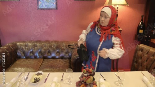 Woman in red shawl on head, Russian traditional clothes stole knife, fruits from served table, put in bag  photo