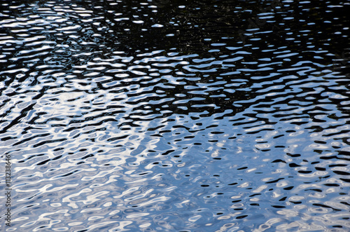 Colourful black, white and blue ripples and reflections of sky, clouds and shadows on water