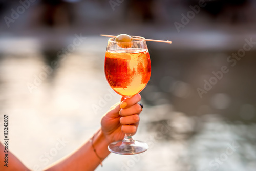 Female hand holding glass with Spritz Aperol alcohol drink decorated with olive on the water chanal background in Venice. Image with small depth of field
