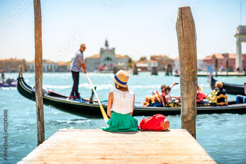 Young female traveler sitting on the pier and enjoying beautiful view on venetian chanal with gondolas floating in Venice © rh2010