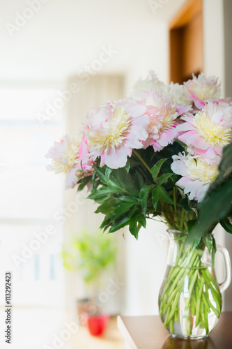 Pretty Bouquet of pink pale peonies in a glass jug with water on the table against the background of a bright room. Home decoration and interior