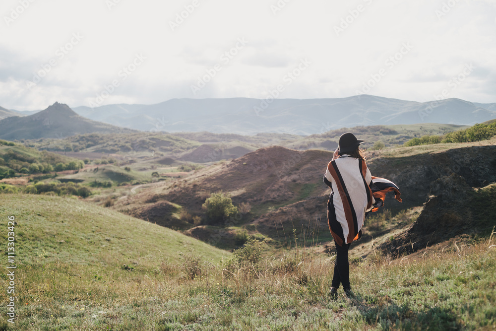 Young beautiful woman traveler wearing hat and poncho relaxing on the top of the hill with mountains and hills around