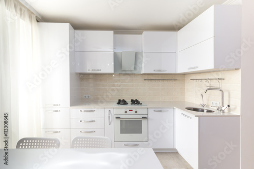 White glossy modern kitchen with stone countertop and built in household appliances in light colors interior