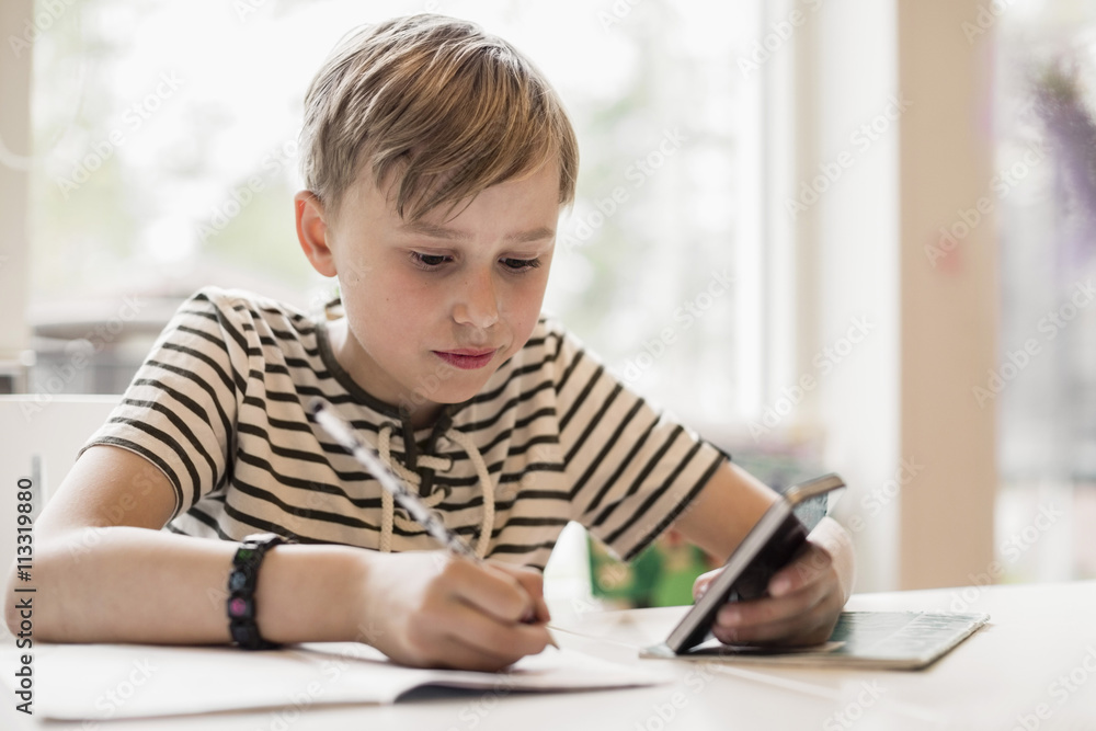 Boy using mobile phone while writing in book at table Boy using mobile phone while writing in book at table