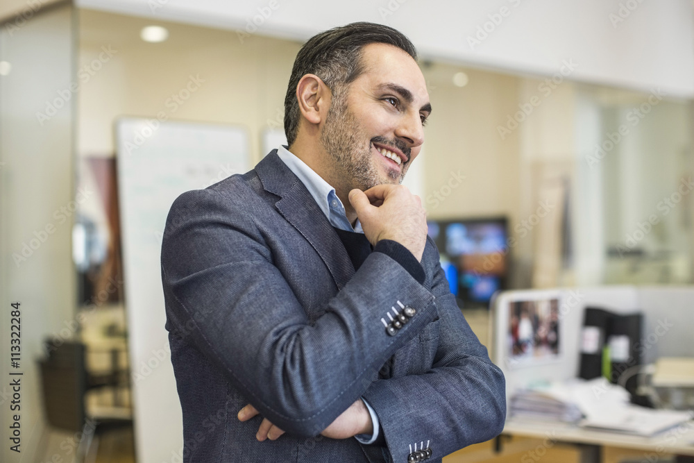 Smiling mid adult businessman with hand on chin looking away in office Smiling mid adult businessman with hand on chin looking away in office