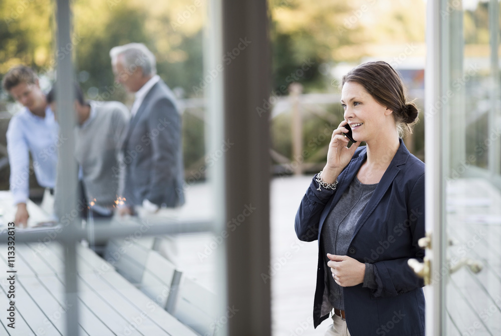 Businesswoman using mobile phone at patio with colleagues working in background Businesswoman using mobile phone at patio with colleagues working in background