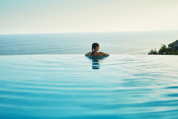 Luxury Resort. Woman Relaxing In Infinity Swimming Pool Water. Beautiful Happy Healthy Female Model Enjoying Summer Travel Vacation, Looking At Sea View. Summertime Recreation, Relax And Spa Concept.