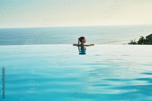 Luxury Resort. Woman Relaxing In Infinity Swimming Pool Water. Beautiful Happy Healthy Female Model Enjoying Summer Travel Vacation, Looking At Sea View. Summertime Recreation, Relax And Spa Concept.