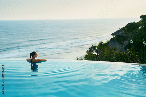 Luxury Resort. Woman Relaxing In Infinity Swimming Pool Water. Beautiful Happy Healthy Female Model Enjoying Summer Travel Vacation, Looking At Sea View. Summertime Recreation, Relax And Spa Concept.