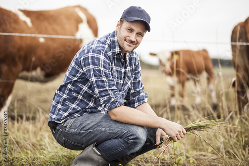 Portrait of farmer with grass crouching on field while animals grazing in background