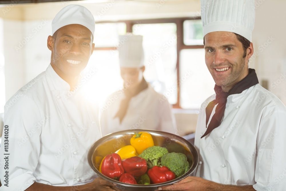 Portrait of two smiling chefs holding a bowl of vegetable Portrait of two smiling chefs holding a bowl of vegetable