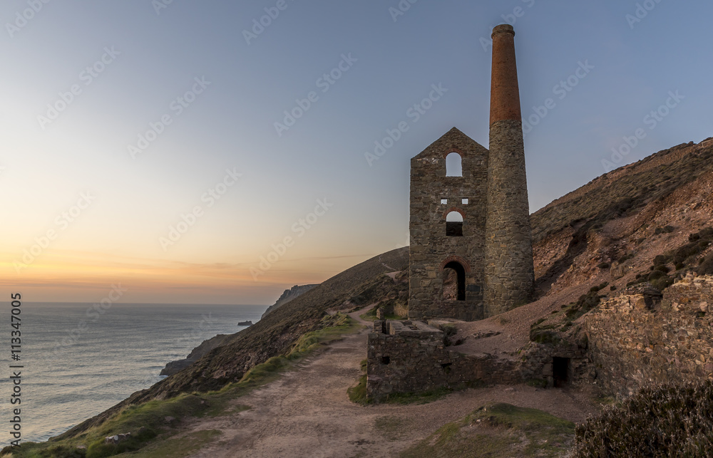 The ruin of a Cornish tin mine, on the edge of a cliff, with the sun setting into the ocean behind it
