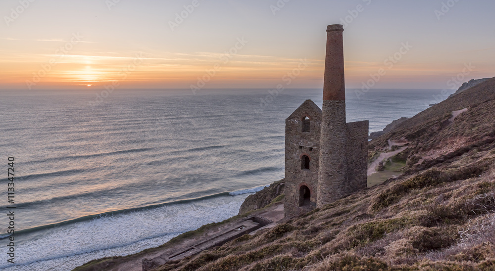 The ruin of a Cornish tin mine, on the edge of a cliff, with the sun setting into the ocean behind it The ruin of a Cornish tin mine, on the edge of a cliff, with the sun setting into the ocean behind it