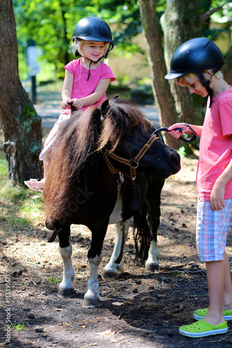 Young children enjoying horse back riding activity. Group of school age kids taking care about their pony and learning how to feed the animal. Summer camp for active holidays.