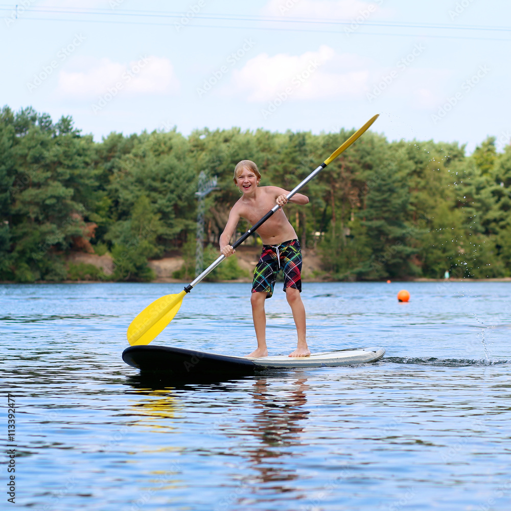 Adventurous boy learning to paddle on stand up board. Happy child, teenage schoolboy, having fun enjoying adventurous experience on the river on a sunny day during summer holidays