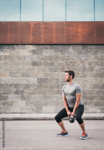 Fitness male working out legs with kettlebell. Sporty man working out outdoor on asphalt.