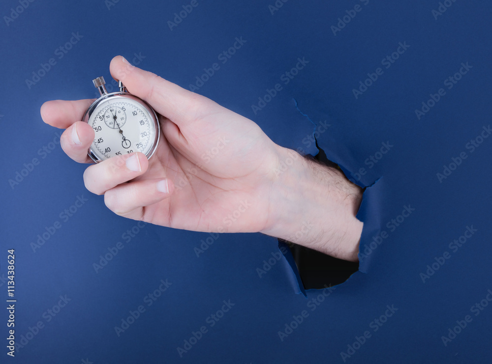 Male hand breaking through the blue paper background and holding antique clock Male hand breaking through the blue paper background and holding antique clock