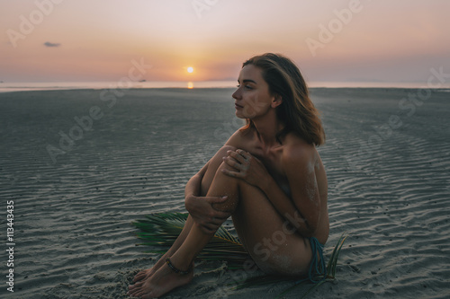 Young pretty girl sitting on the sand during sunset with palm branch