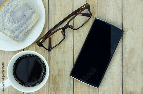 Smartphone, eyeglasses and a cup of coffee with bread on wooden