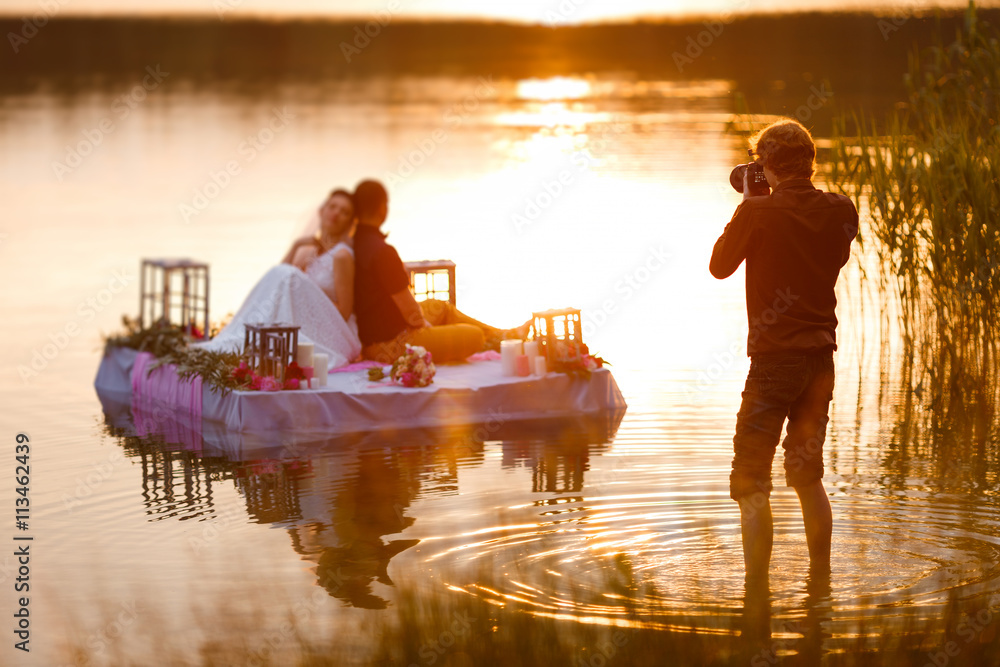 Wedding photographer in action, taking a picture of the bride and groom sitting on the raft. Summer, sunset - obrazy, fototapety, plakaty 
