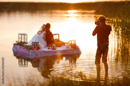 Wedding photographer in action, taking a picture of the bride and groom sitting on the raft. Summer, sunset