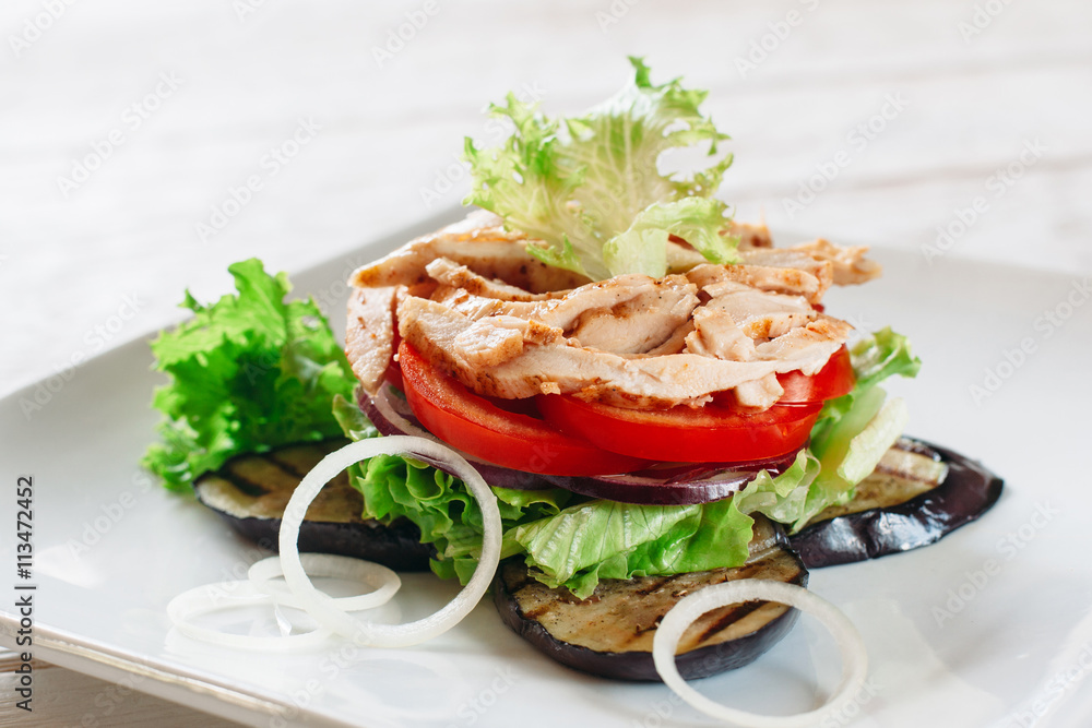 Layered salad with chicken, tomato and grilled eggplant on white. White square plate with portion of layered salad with fried chicken strips, tomato, lettuce, onion and grilled eggplant.