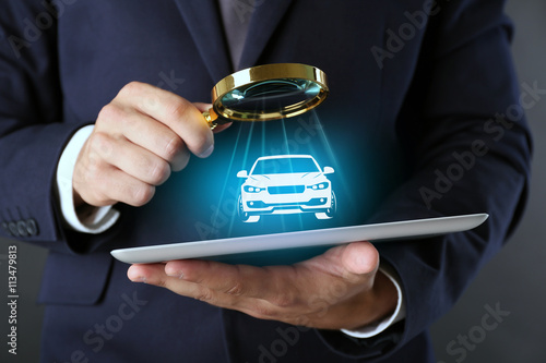 Businessman holding magnifying glass and digital tablet with virtual car on dark grey background