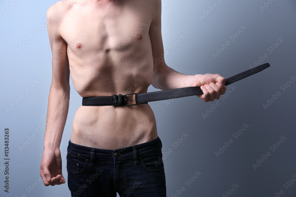 Skinny young man with anorexia tightening his waist with belt on grey  background Stock Photo