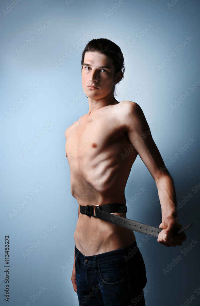 Skinny young man with anorexia tightening his waist with belt on grey  background Stock Photo