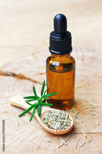a bottle of rosemary aromatherapy oil extract with fresh and dried rosemary leaf on wooden spoon. 