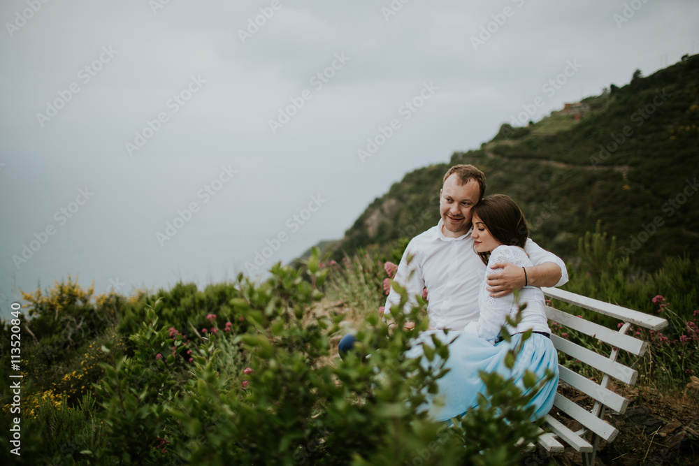 Young cute couple honeymoon on a white chair holding their hands on dating in a beautiful place italy near ocean and mountains, hug, smile and talk to each other

