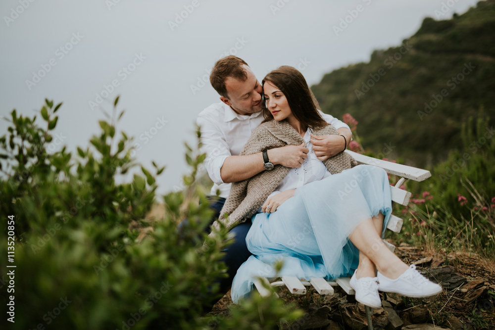 Fototapeta premium Young cute couple honeymoon on a white chair holding their hands on dating in a beautiful place italy near ocean and mountains, hug, smile and talk to each other 