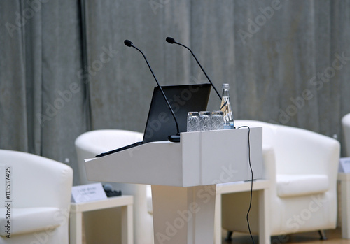 Белая кафедра для выступления с двумя микрофонами, открытым ноутбуком и стаканами на фоне кресел президиума на сцене