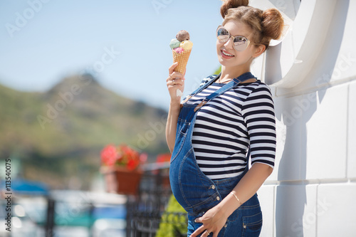 Beautiful pregnant brunette woman with trendy hairstyle,beautiful smile,dressed in blue denim overalls and striped t-shirt,mirrored sunglasses,posing on the promenade with a cone of colored ice cream © GTeam
