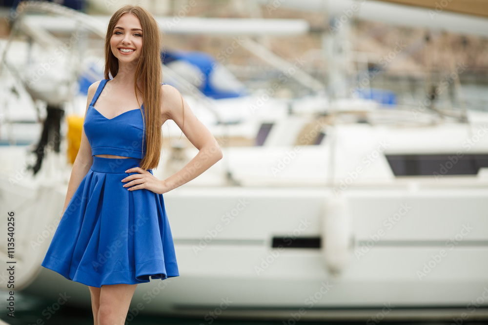 Slender blond young woman with gray-green eyes and long straight hair, dressed in a blue sundress,a beautiful smile posing outdoors in the summer against white yachts and boats on the dock