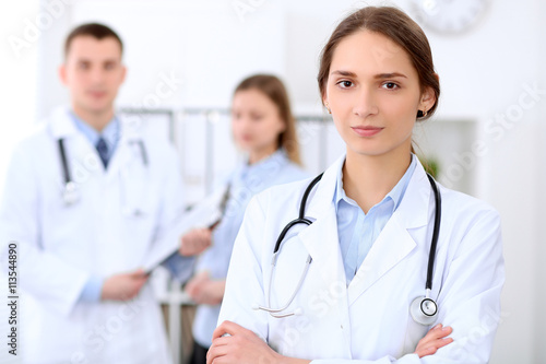 Young beautiful female doctor smiling  on the background with patient and his doctor in hospital. High level and quality medical service concept.