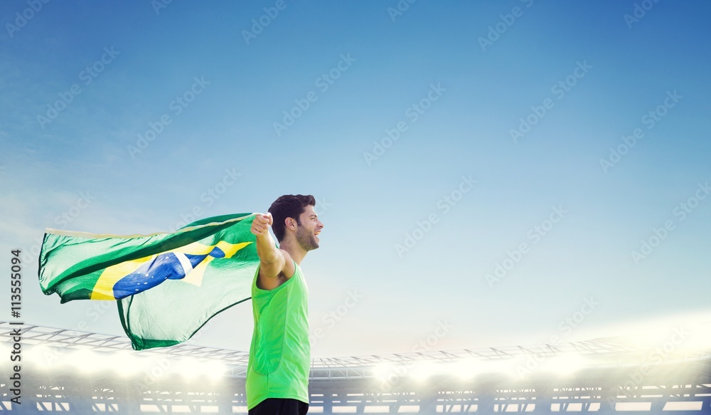 Profile view of Brazilian sportsman is holding a flag against large football stadium under bright blue sky Profile view of Brazilian sportsman is holding a flag against large football stadium under bright blue sky
