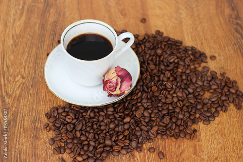 White Coffee mug with saucer and dried red rose and coffee beans on a wooden background closeup