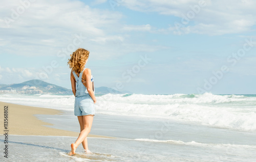 Young woman in blue denim jumpsuit walking along beach and the stormy ocean on sunny day