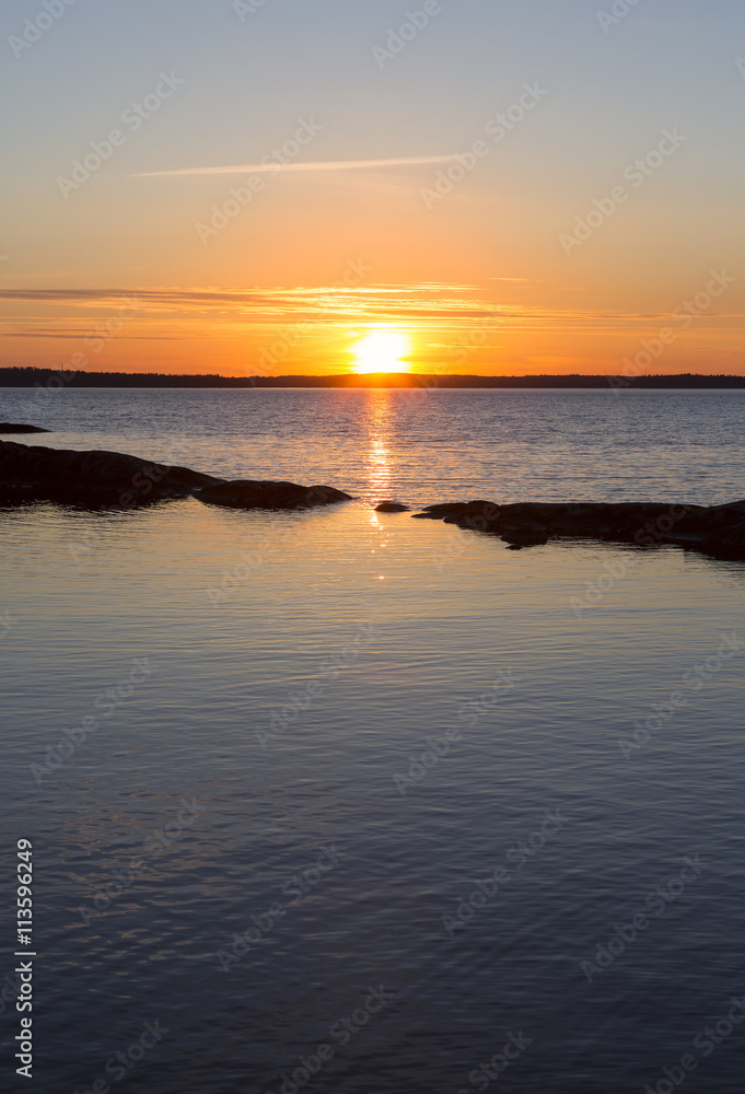 Sun is going down far away on a peaceful night. Some stones are like small island in the front. Glowing sky is colorful in the evening in Finland.