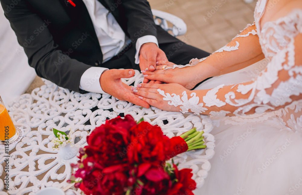 Hands of a newly-married couple close up with flowers boquet on white stylish table. Hands of a newly-married couple close up with flowers boquet on white stylish table.