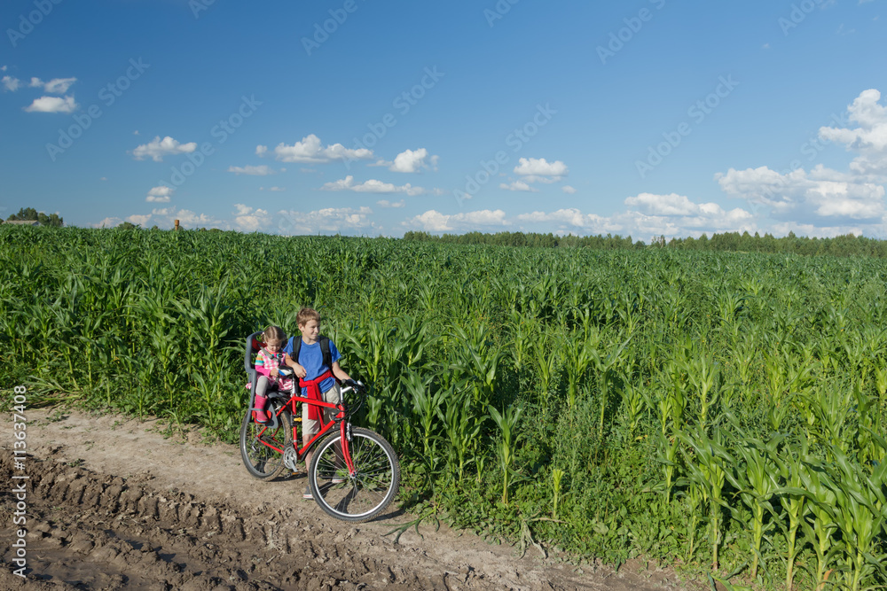 Obraz premium Teenage boy carrying his little sibling sister on baby bike seat on farm corn field summer dirt road