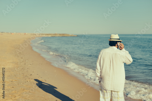 Person talking on cell phone walking by open water blue sky horizon, sunlight outdoors.