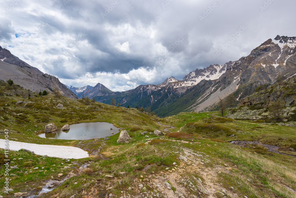 High altitude alpine pond in extrem terrain rocky landscape once covered by glaciers. Dramatic stormy sky and snowcapped mountain range, Italian French Alps.